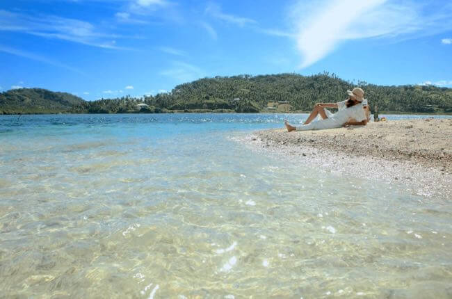 “Serene tropical beach scene in Fiji featuring a woman relaxing on soft white sand by clear turquoise waters under a bright,